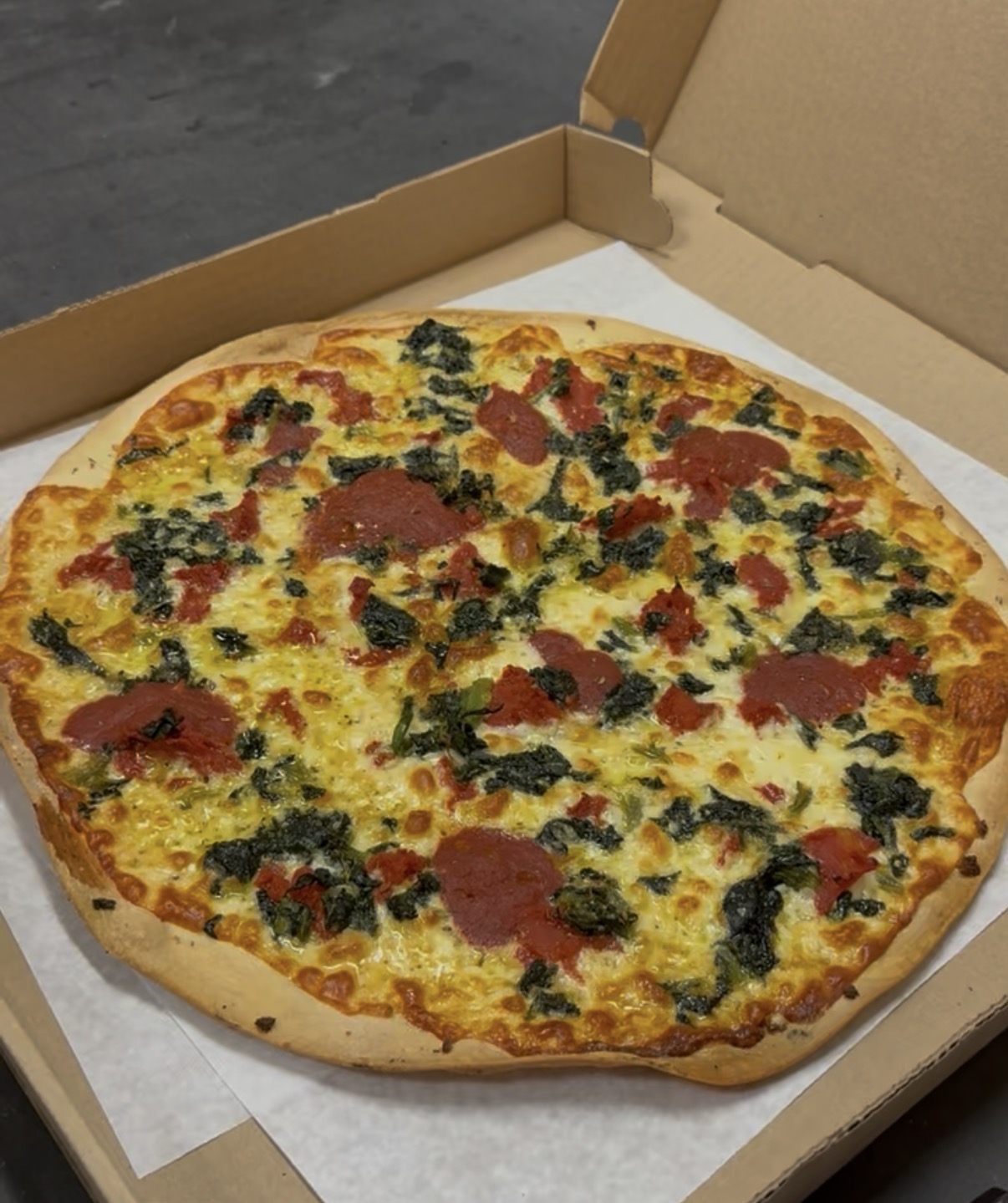 Cheese pizza with pepperoni and spinach in an open cardboard box on white paper.