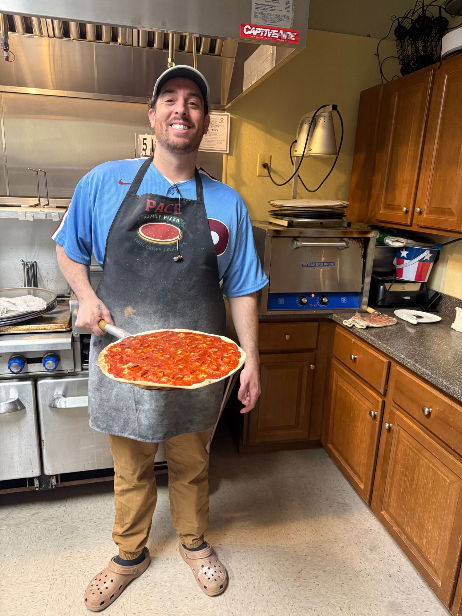 Man in apron holding pizza in kitchen with wooden cabinets and stainless steel appliances.