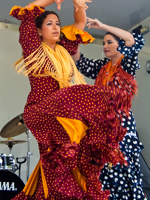 Two women in colorful polka dot dresses perform a dance indoors, with musical instruments visible in the background.