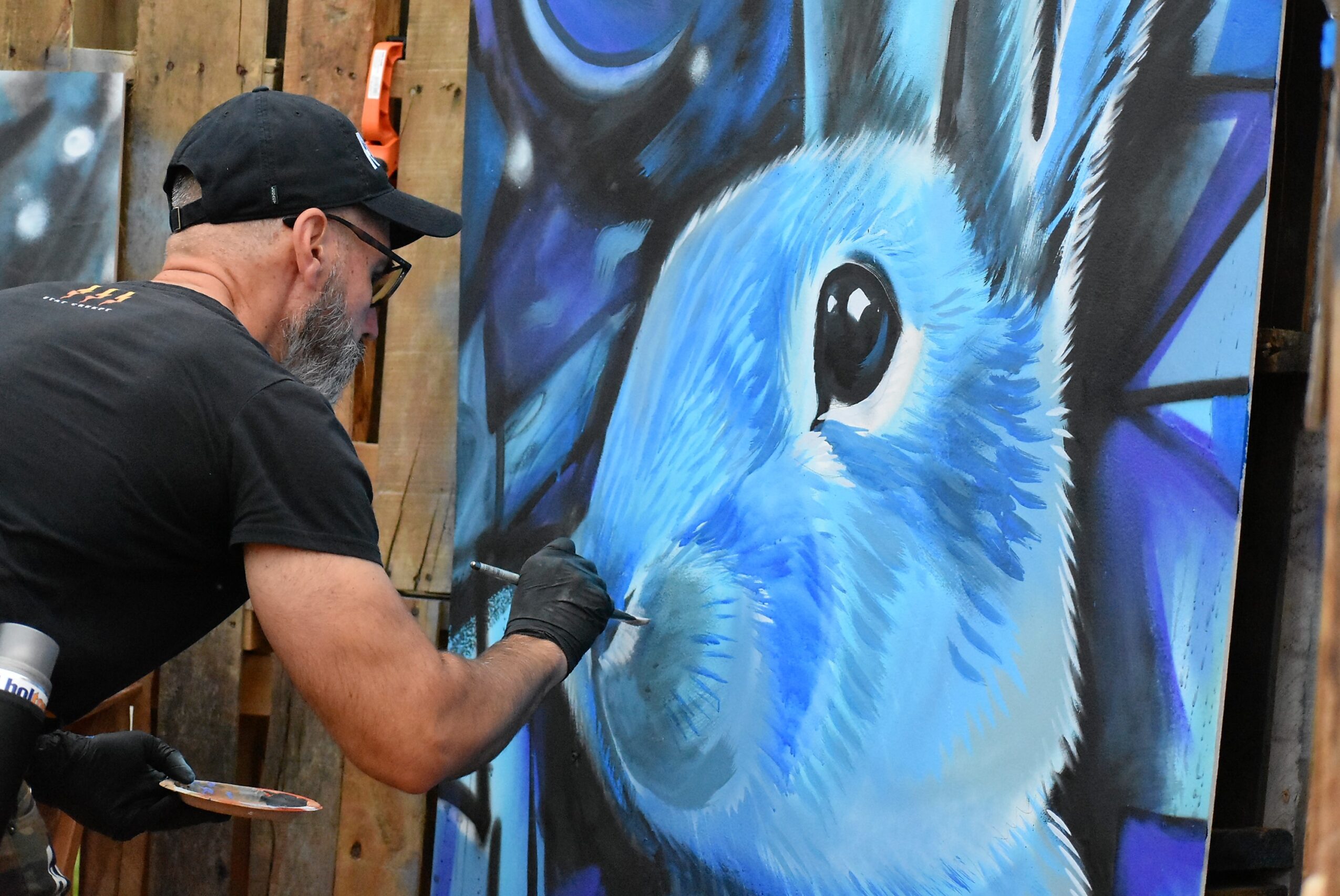 Artist painting a blue rabbit on a large canvas with a brush, wearing a black cap and gloves.