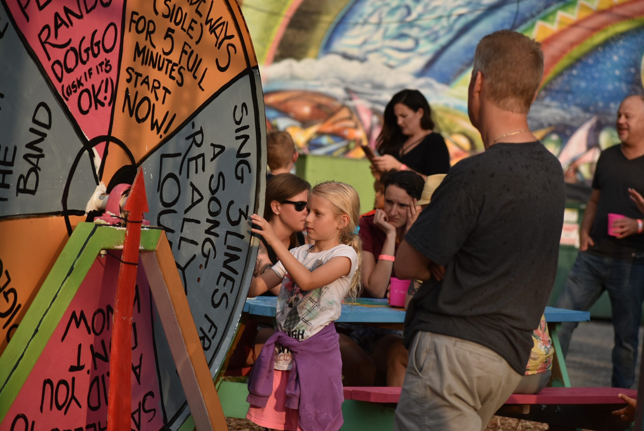 Children and adults gather around a colorful spinning wheel at an outdoor event, with a rainbow backdrop and picnic tables.