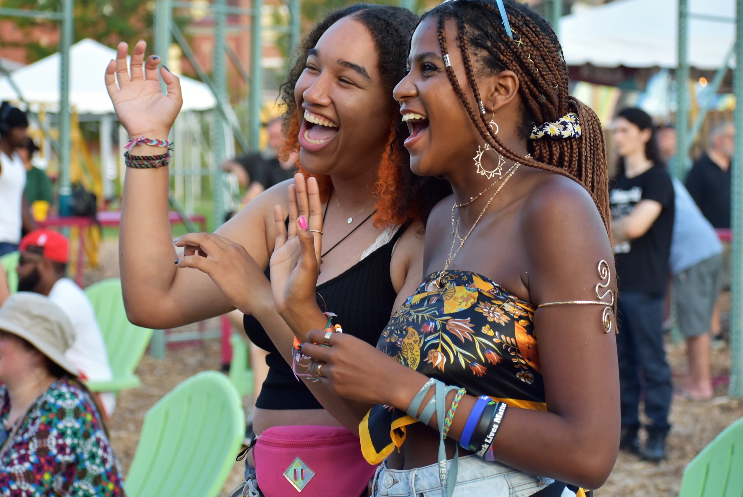 Two women laughing and talking outdoors at a festival or fair, with people and tents in the background.