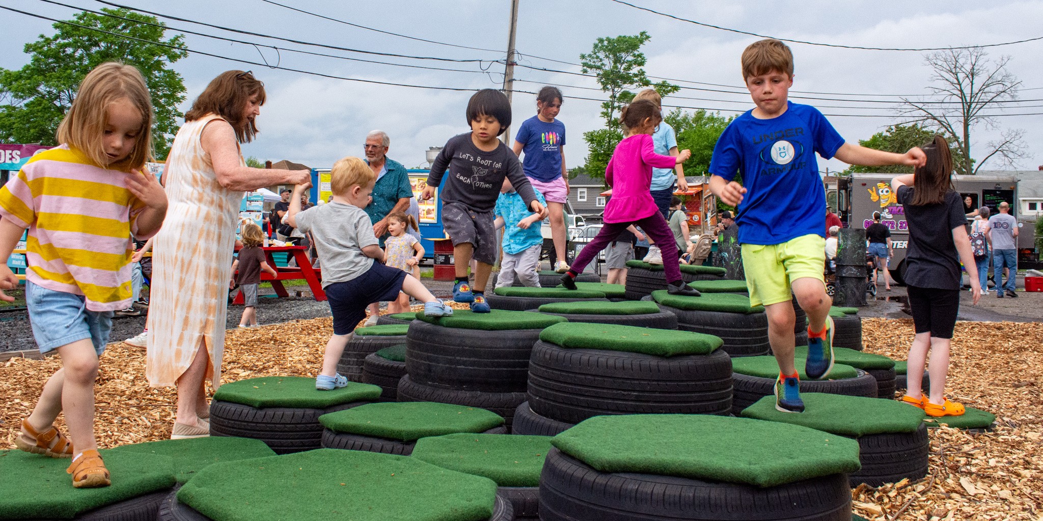Children playing on a tire obstacle course outdoors with trees and a cloudy sky in the background.