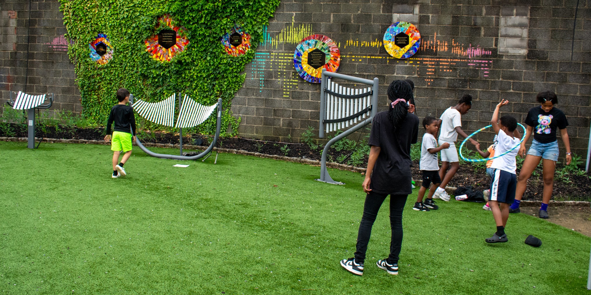 Children and adults playing on a grassy area with colorful flower decorations on a brick wall.