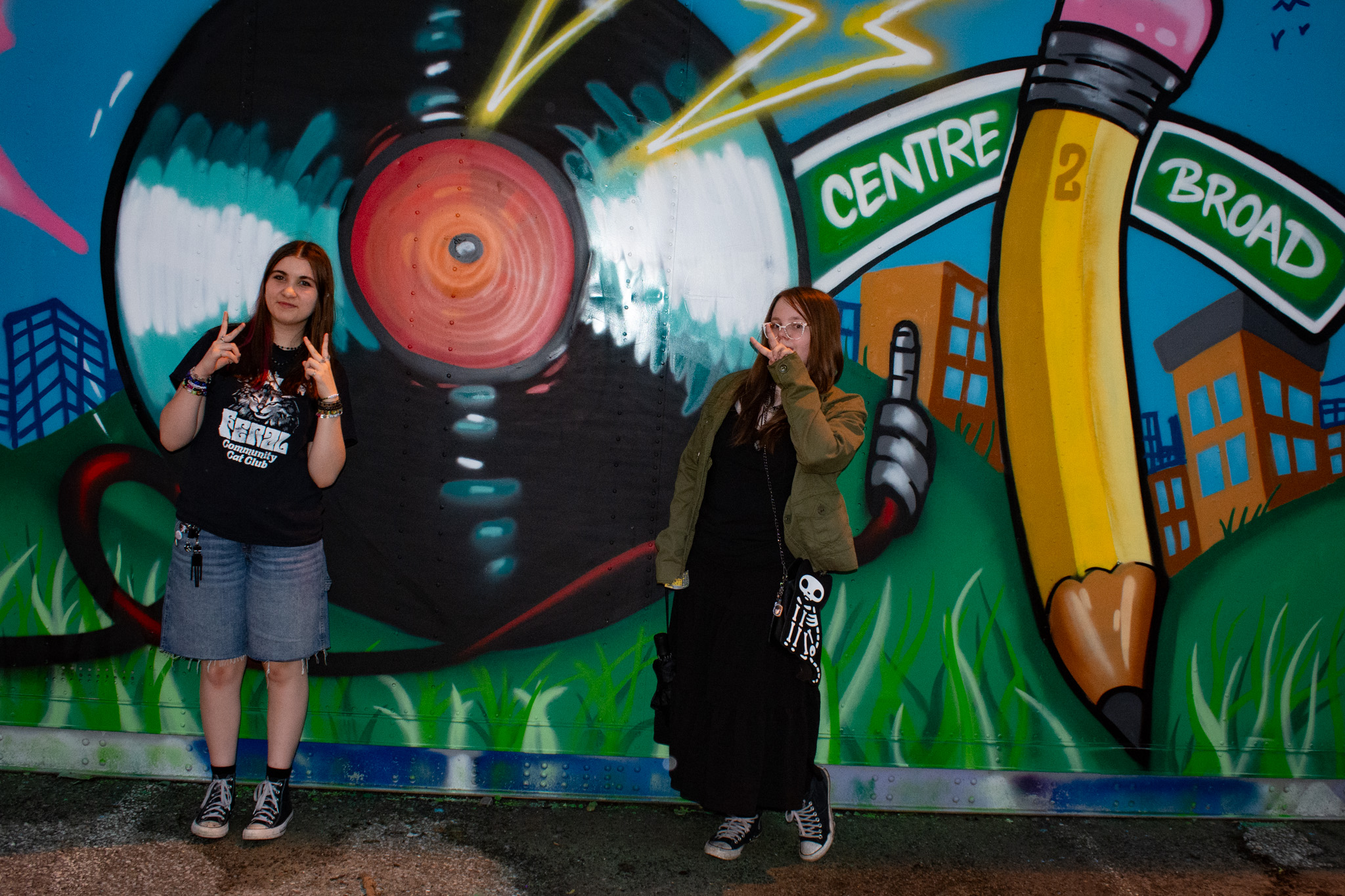 Two women stand in front of a colorful graffiti mural with a large eye and cityscape.