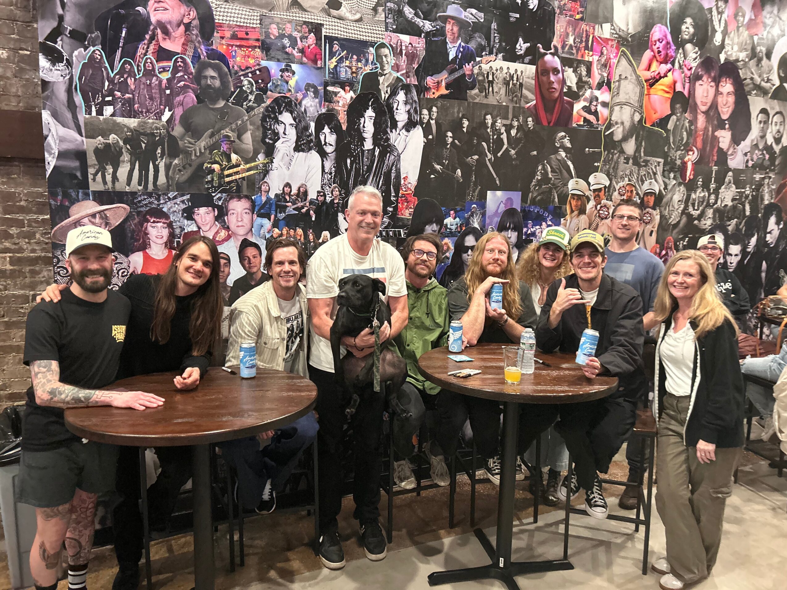 Group of people gathered around two tables, smiling, with a collage of photos on the wall behind them.