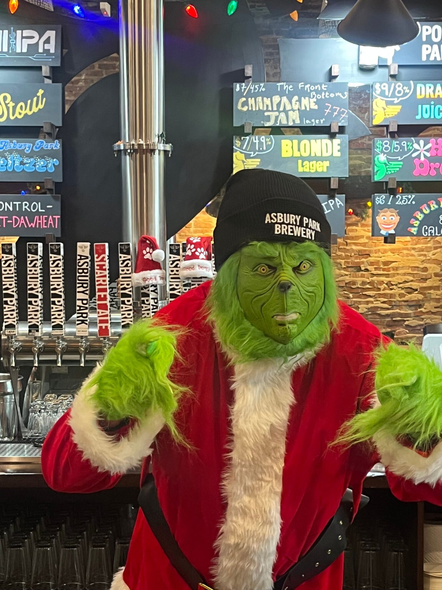 Person dressed as a green creature with a face resembling the Grinch, wearing a Santa suit and a black hat, in a bar setting.