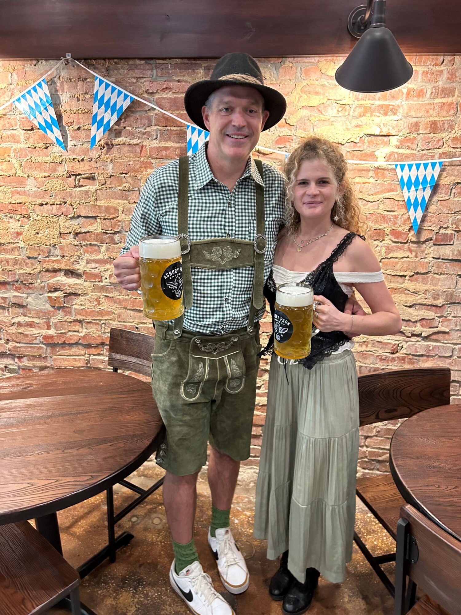 Man and woman in traditional Bavarian attire holding beer mugs, standing in a decorated indoor setting with brick wall and blue flags.
