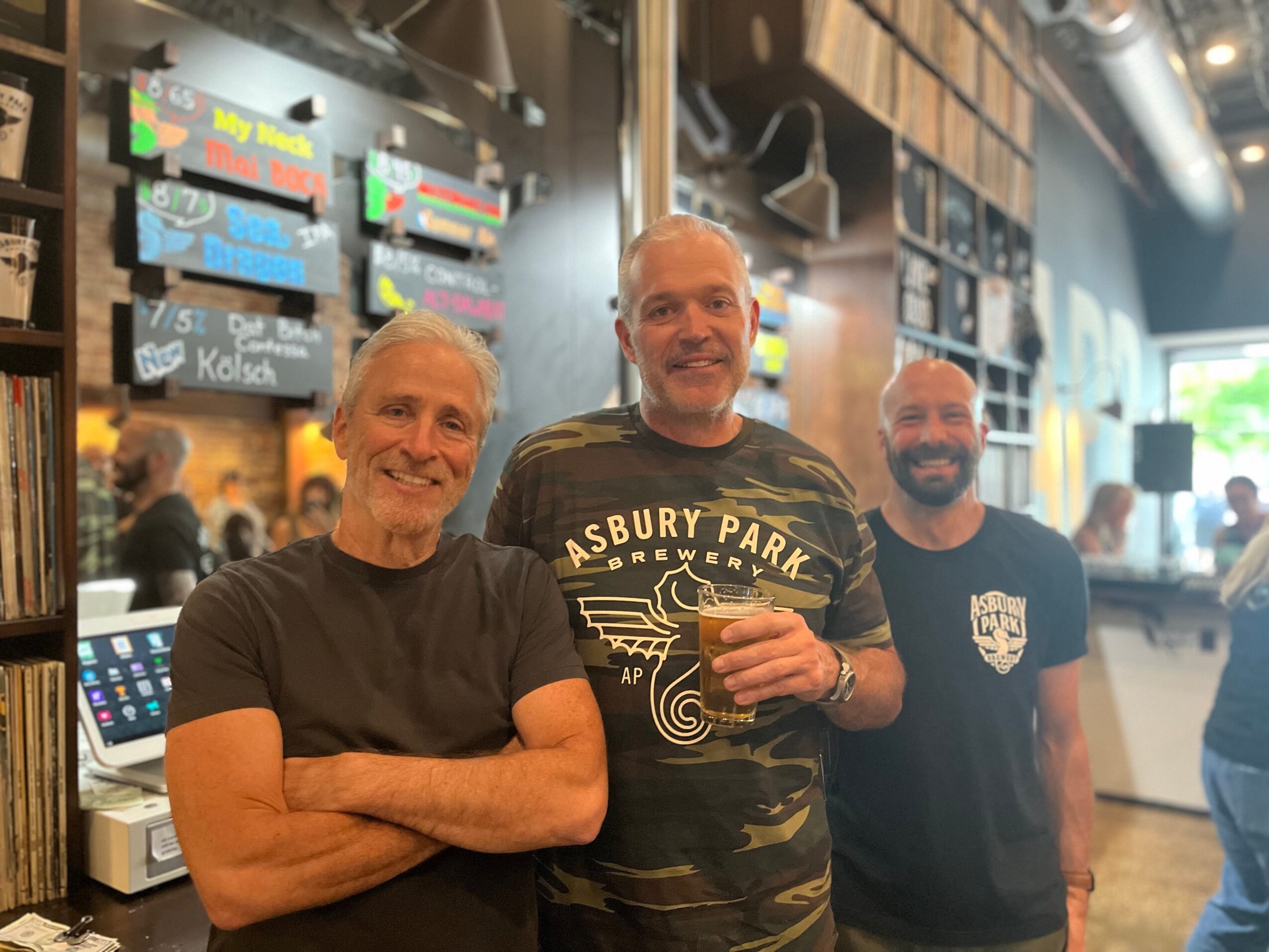 Three men standing inside a cafe, smiling, with menu boards and a window in the background.