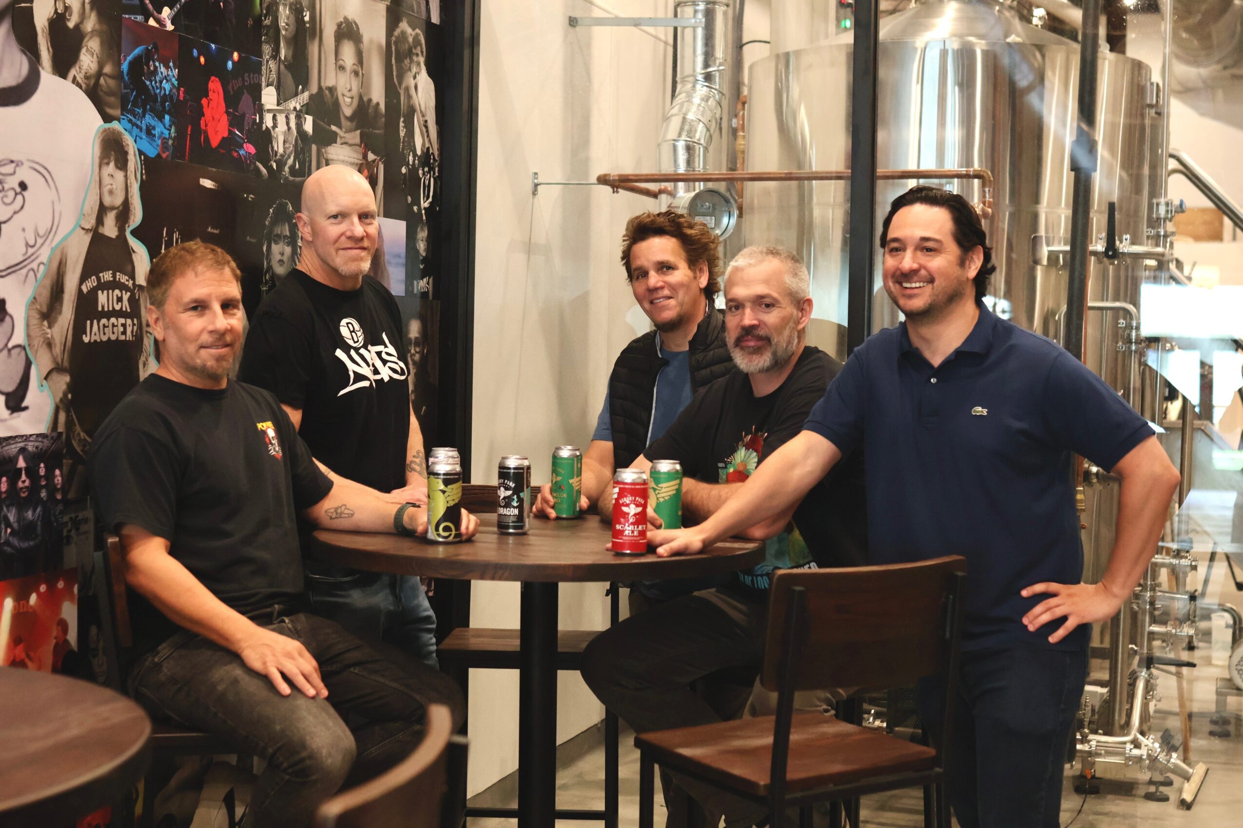 Five men gathered around a table with drinks in a brewery setting, smiling and posing for the photo.