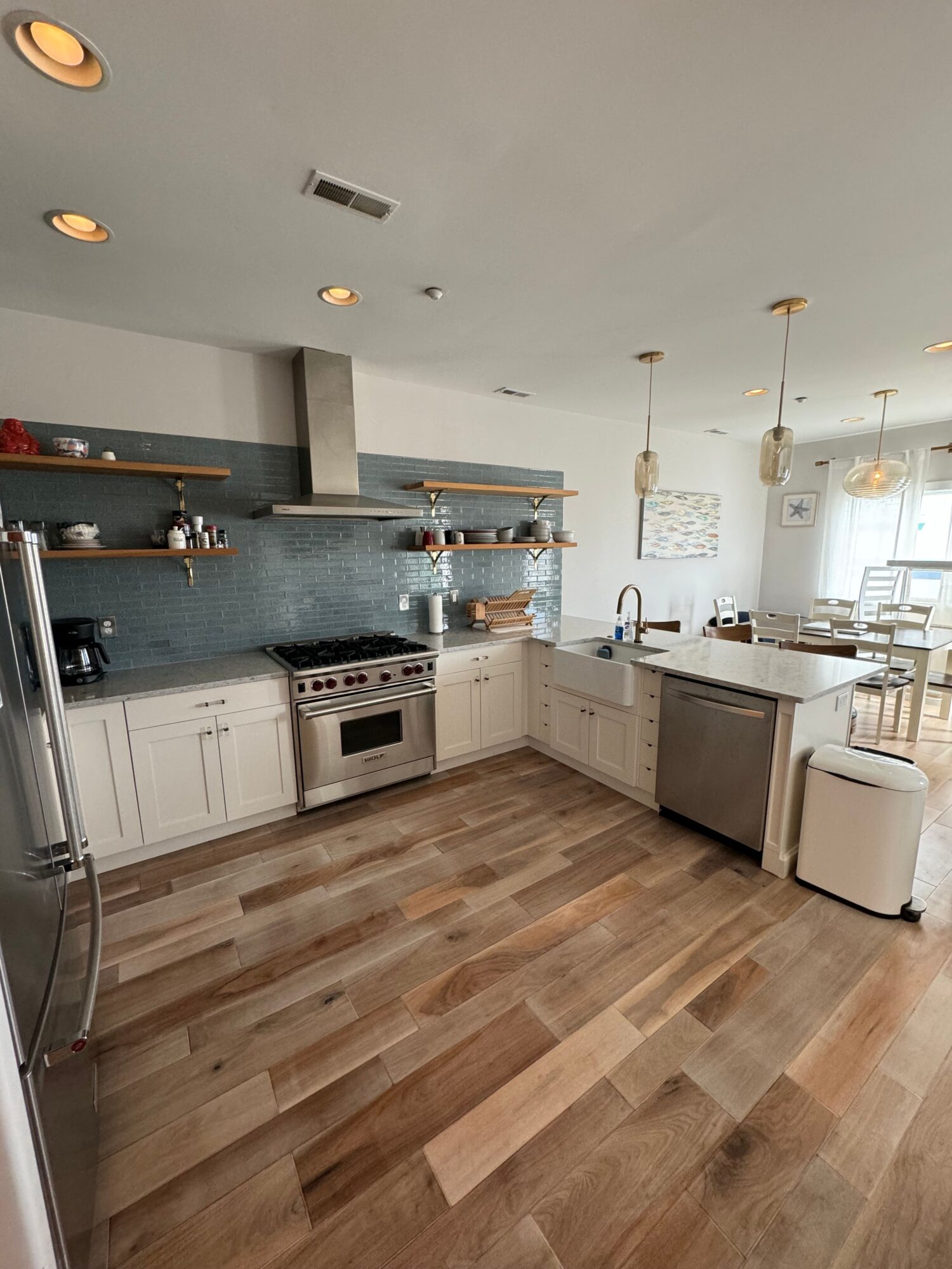 Modern kitchen with white cabinets, open shelves, stainless steel appliances, and a blue backsplash, illuminated by pendant lights and natural light.