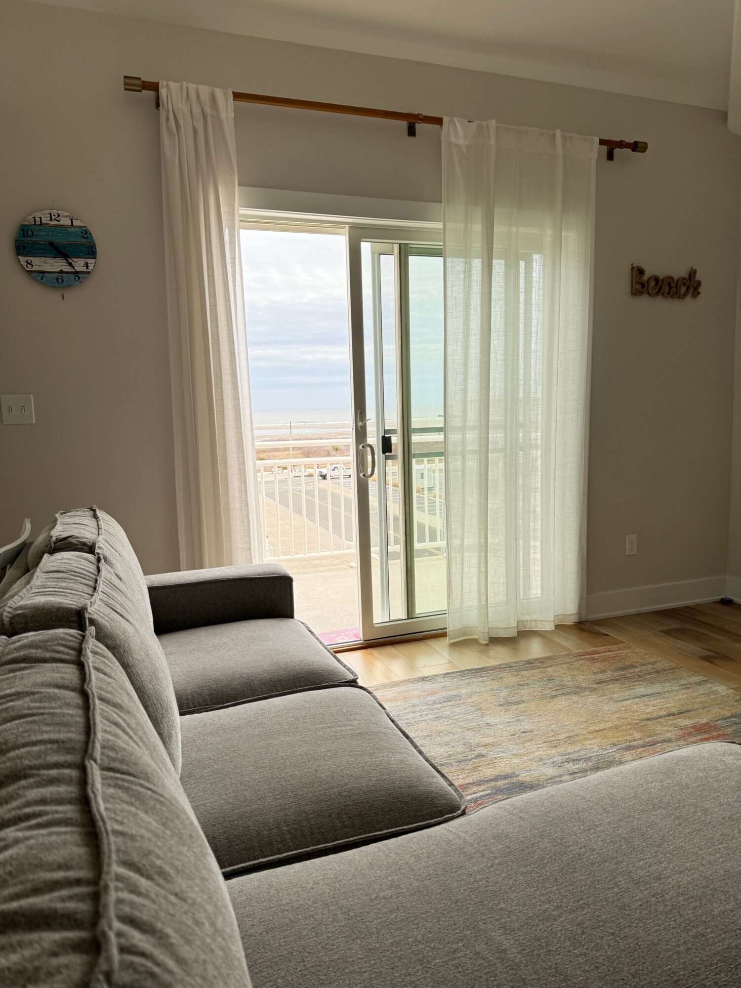 Living room with sliding glass door, sheer curtains, gray sofa, wooden floor, and beach view outside.
