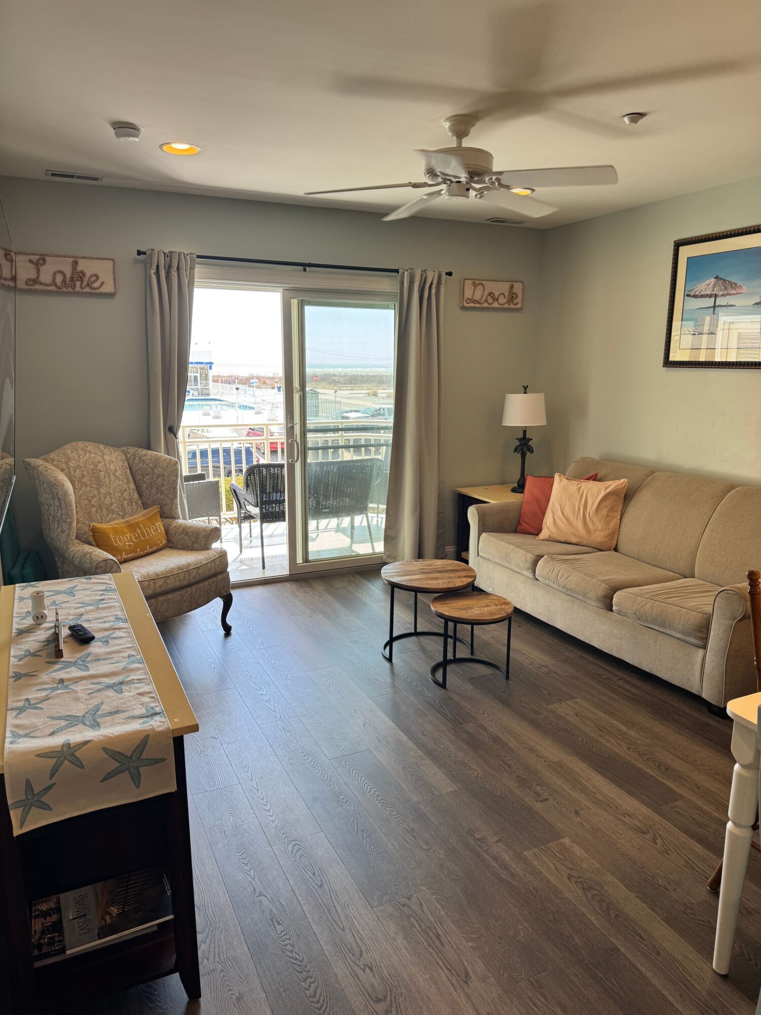 Living room with sofa, armchair, sliding glass door, and ocean view, decorated with beach-themed signs and a lamp.