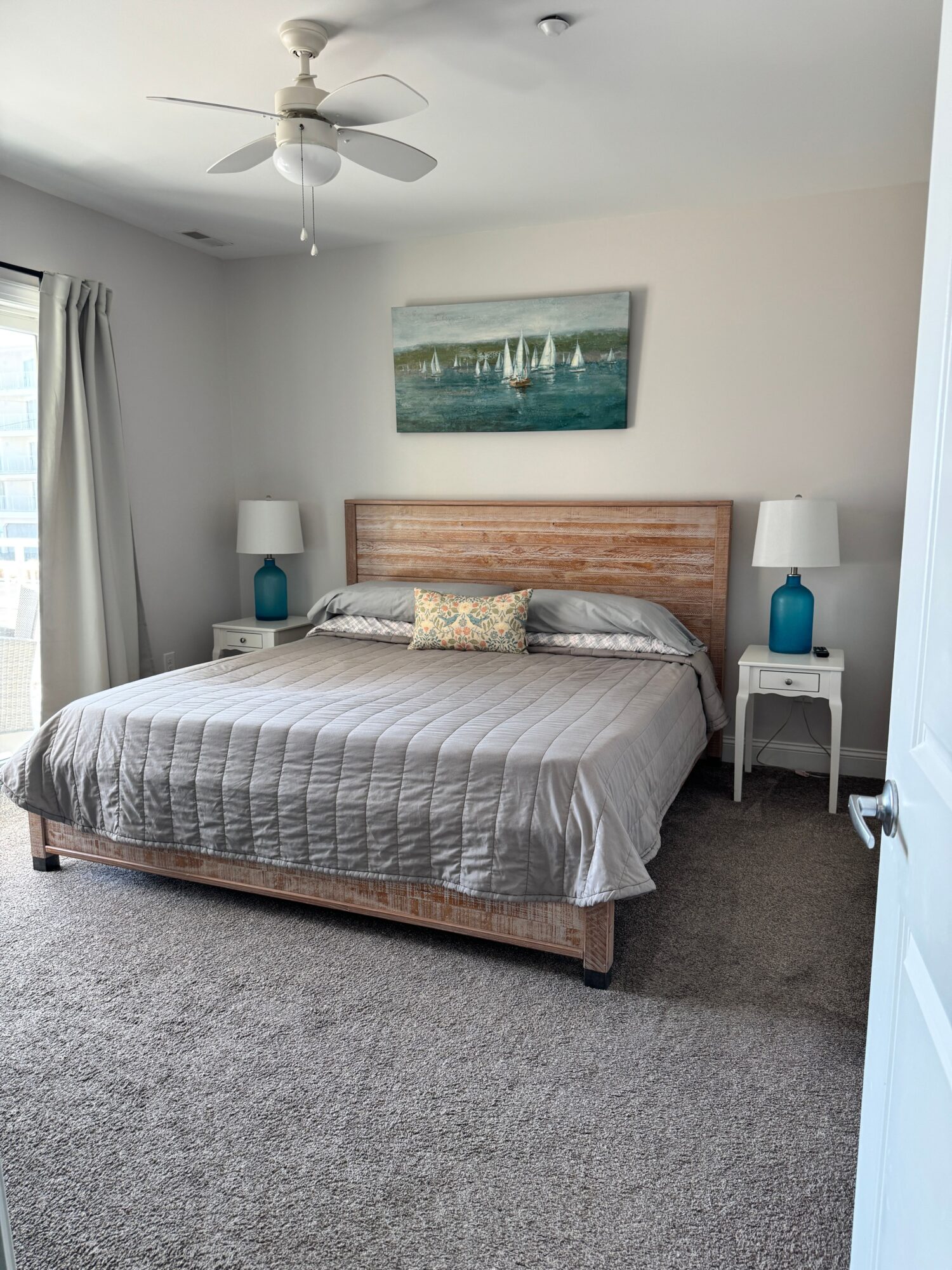 Bedroom with bed, two nightstands with lamps, painting above headboard, window with curtains, ceiling fan, gray carpet.