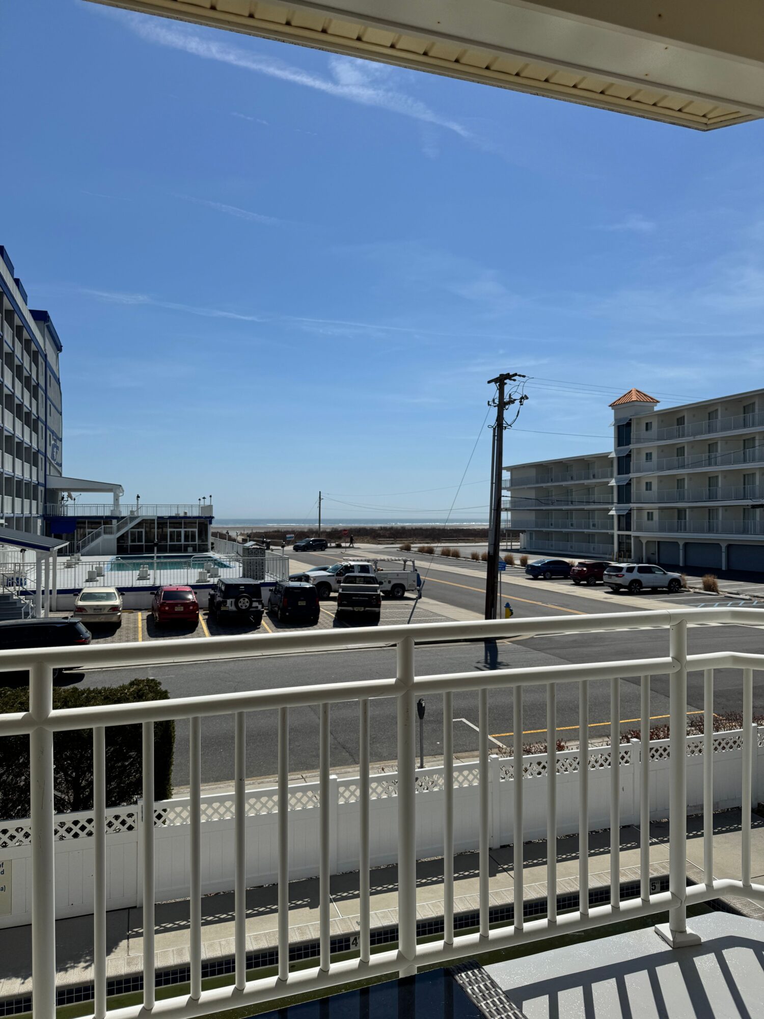 View of a parking lot with cars, buildings, and a clear blue sky, seen from a balcony with white railing.