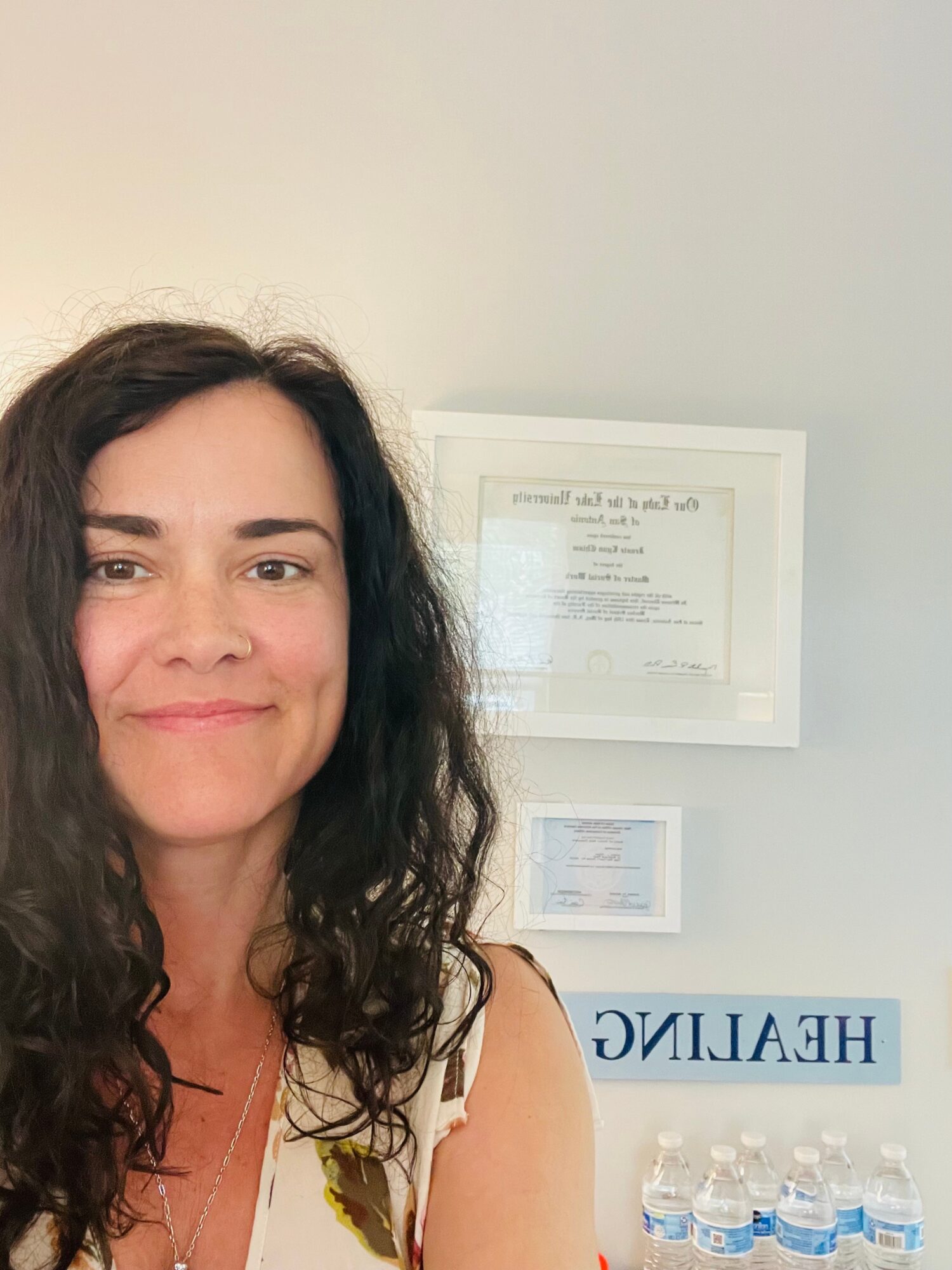 Woman with long dark curly hair smiling, framed certificates on white wall behind her, HELALIC sign visible.