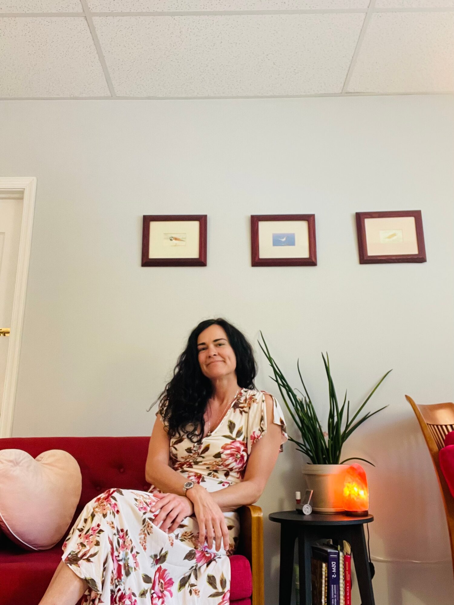 Woman with long dark hair sitting on a red sofa, smiling, wearing a floral dress, next to a large plant and a candle.