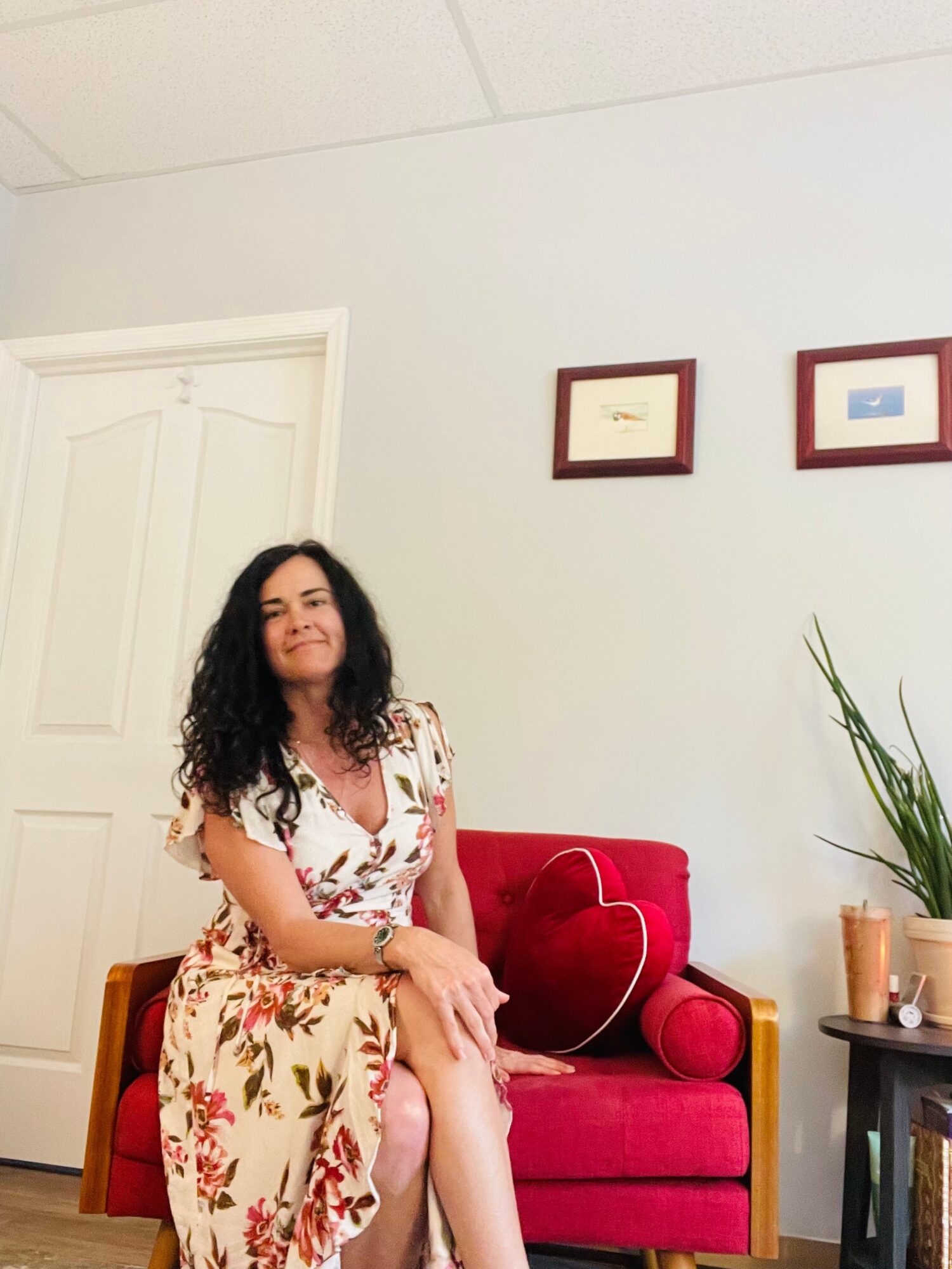 Woman with curly hair sitting on a red velvet sofa in a room with framed pictures and a plant.