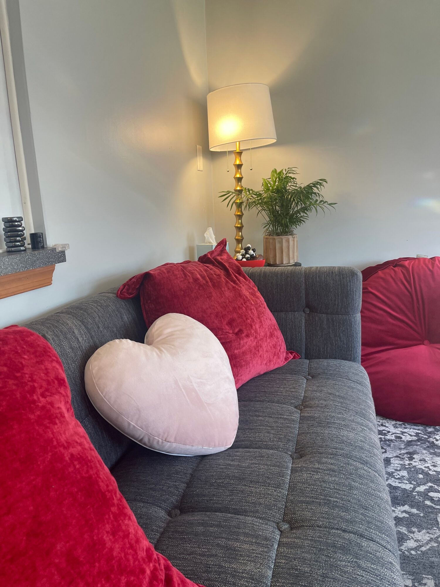 Gray sofa with red and white heart-shaped pillows, a lamp, and a potted plant on a side table.