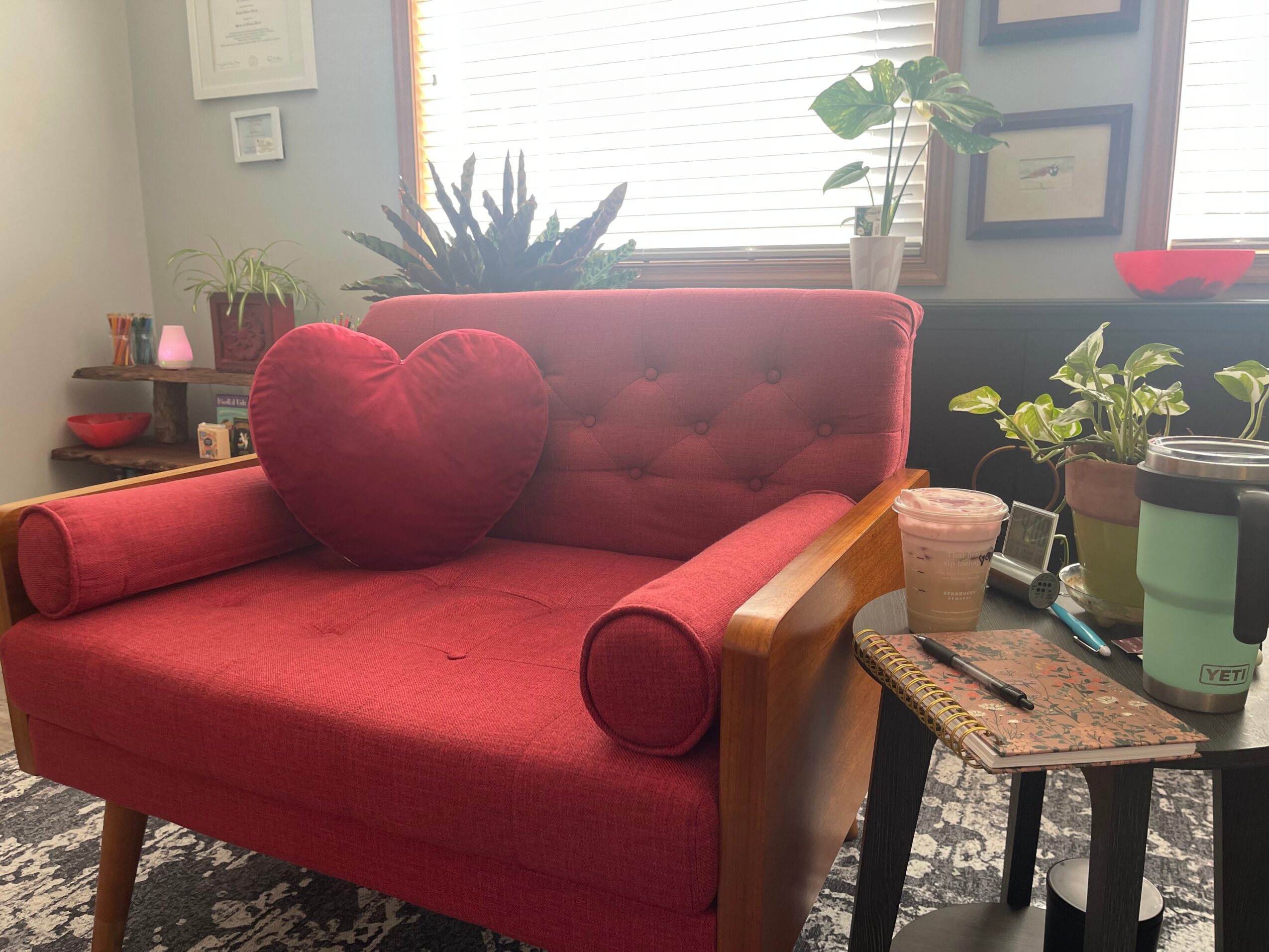 Red armchair with a heart-shaped pillow, wooden armrests, in a room with plants and framed pictures on the wall.