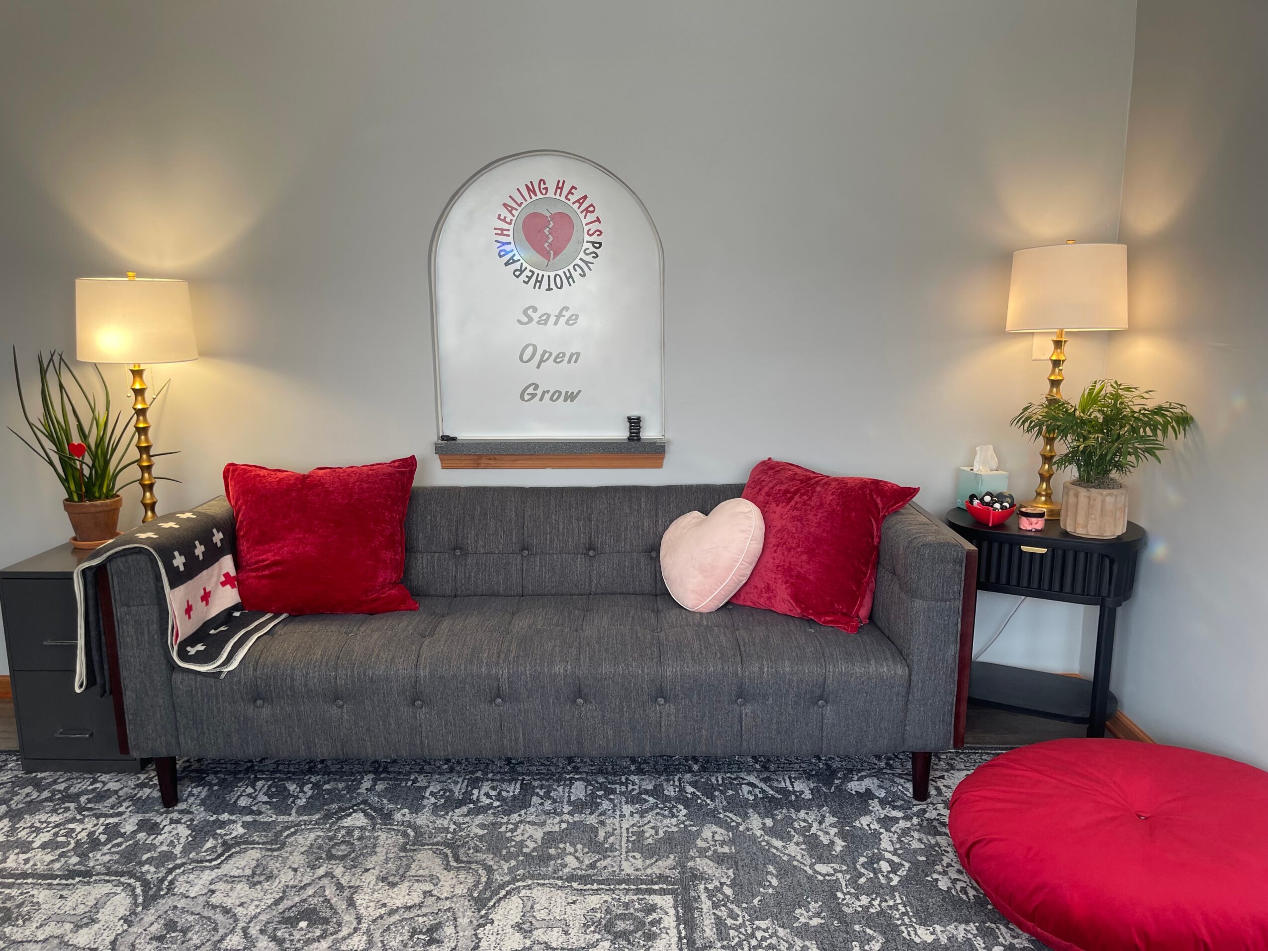 Gray sofa with red and pink pillows, two lamps, a black side table with plants, and a red cushion on the floor.