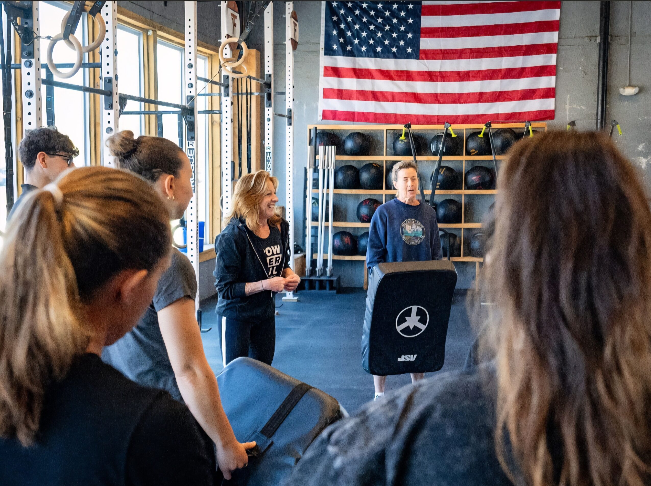 Group of people listening to a woman speaking in a gym with an American flag and fitness equipment in the background.