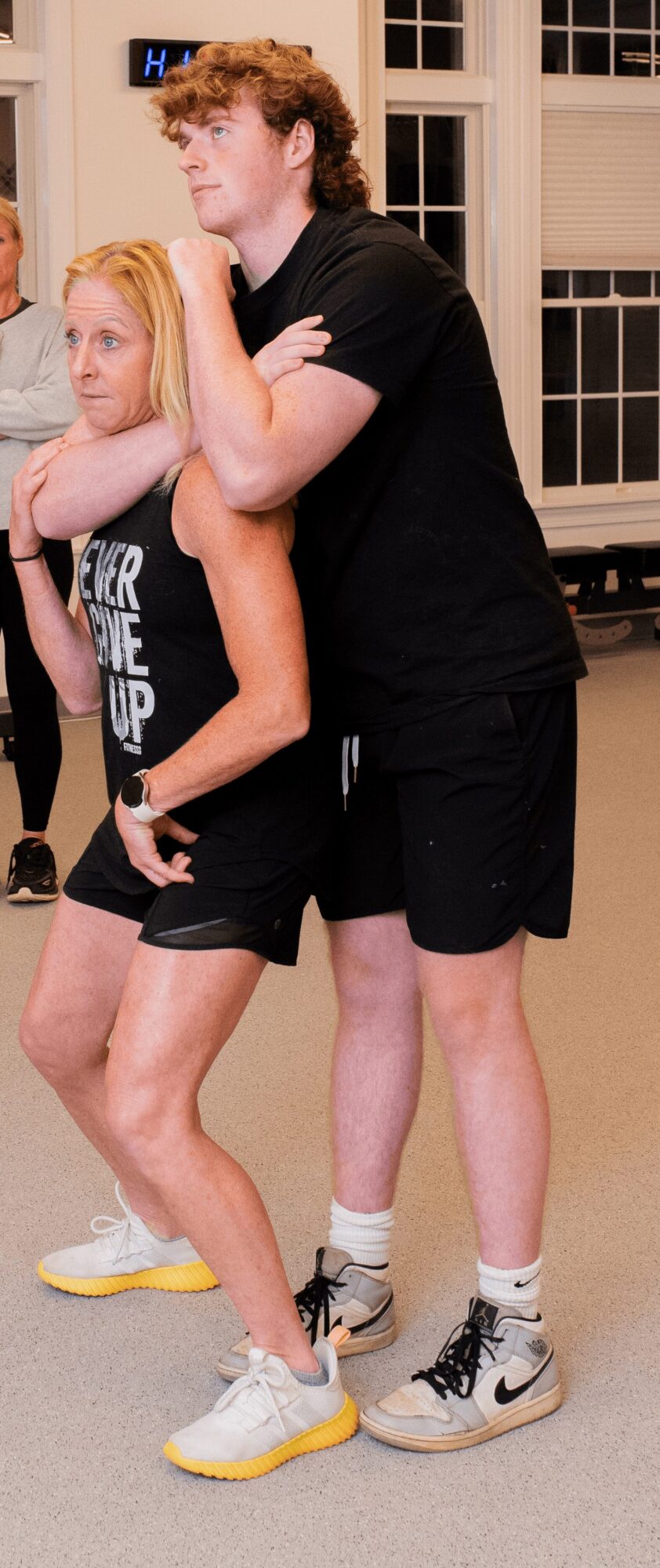 Two people in a gym or sports facility, one person standing behind and holding the other from behind.