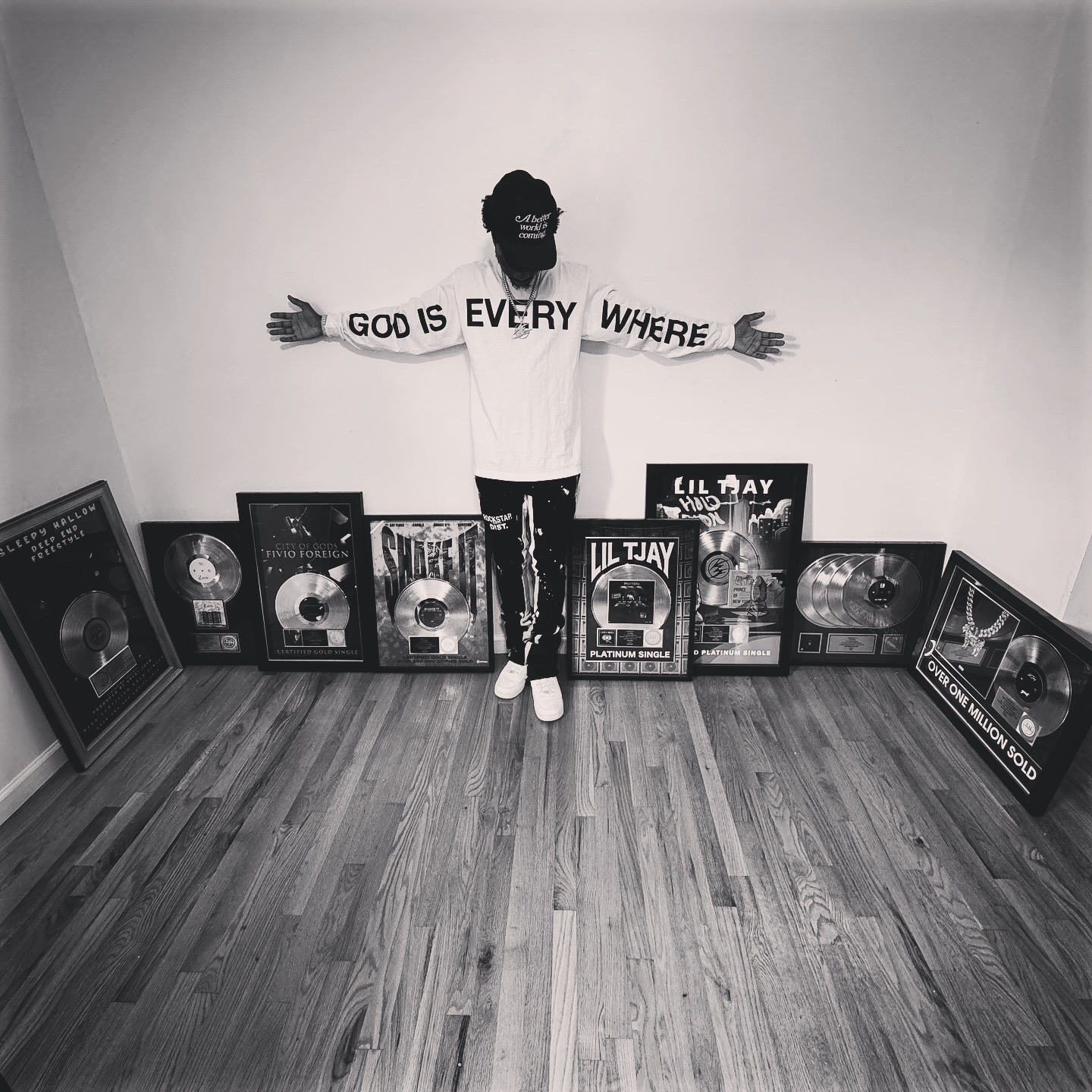 Person standing with arms outstretched, holding a sign that says 'GOD IS EVERY WHERE', surrounded by framed music awards on the floor.