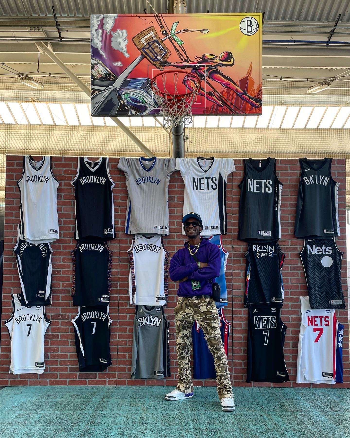 Person standing in front of basketball jerseys and a colorful mural of a basketball game, wearing sunglasses, a blue jacket, and camouflage pants.