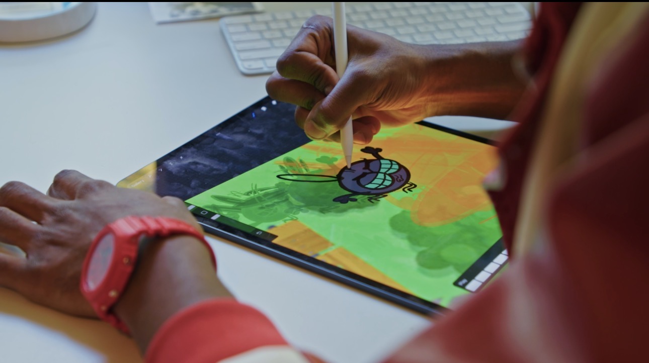 Person drawing on a tablet with a stylus, colorful cartoon character on screen, workspace with keyboard and phone nearby.