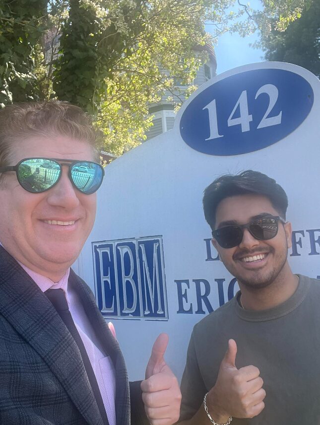 Two men wearing sunglasses smiling and giving thumbs up outdoors with a sign displaying the number 142 and logos behind them.