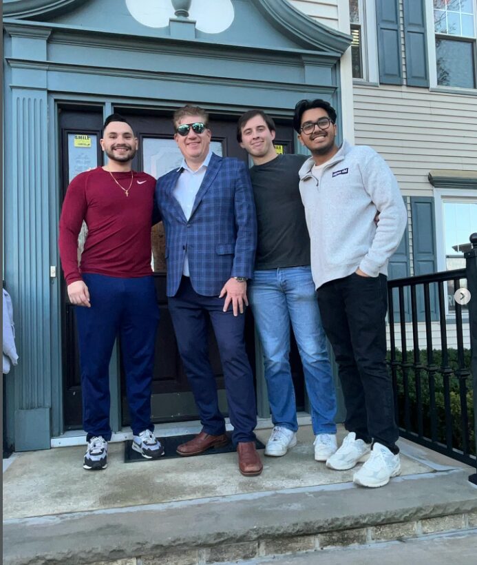 Four men standing outside a building, smiling, with a railing and windows behind them.
