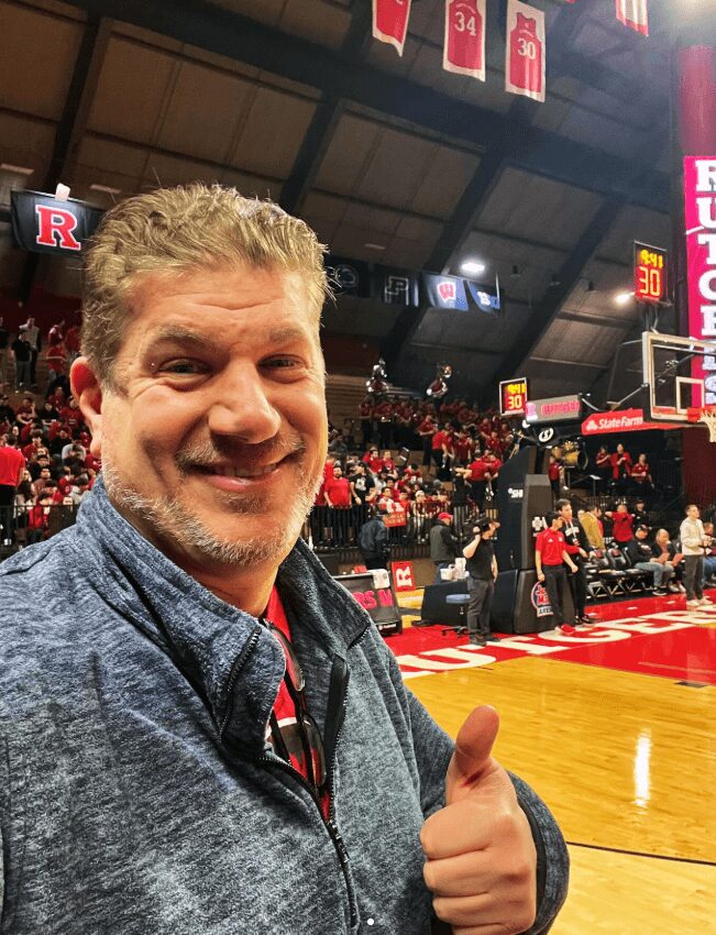 Smiling man giving a thumbs-up at a basketball game, with a court and spectators in the background.