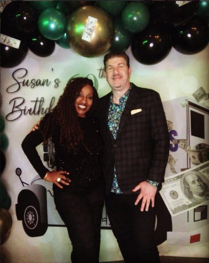 Two people smiling at a birthday celebration, decorated with balloons and a money-themed backdrop.