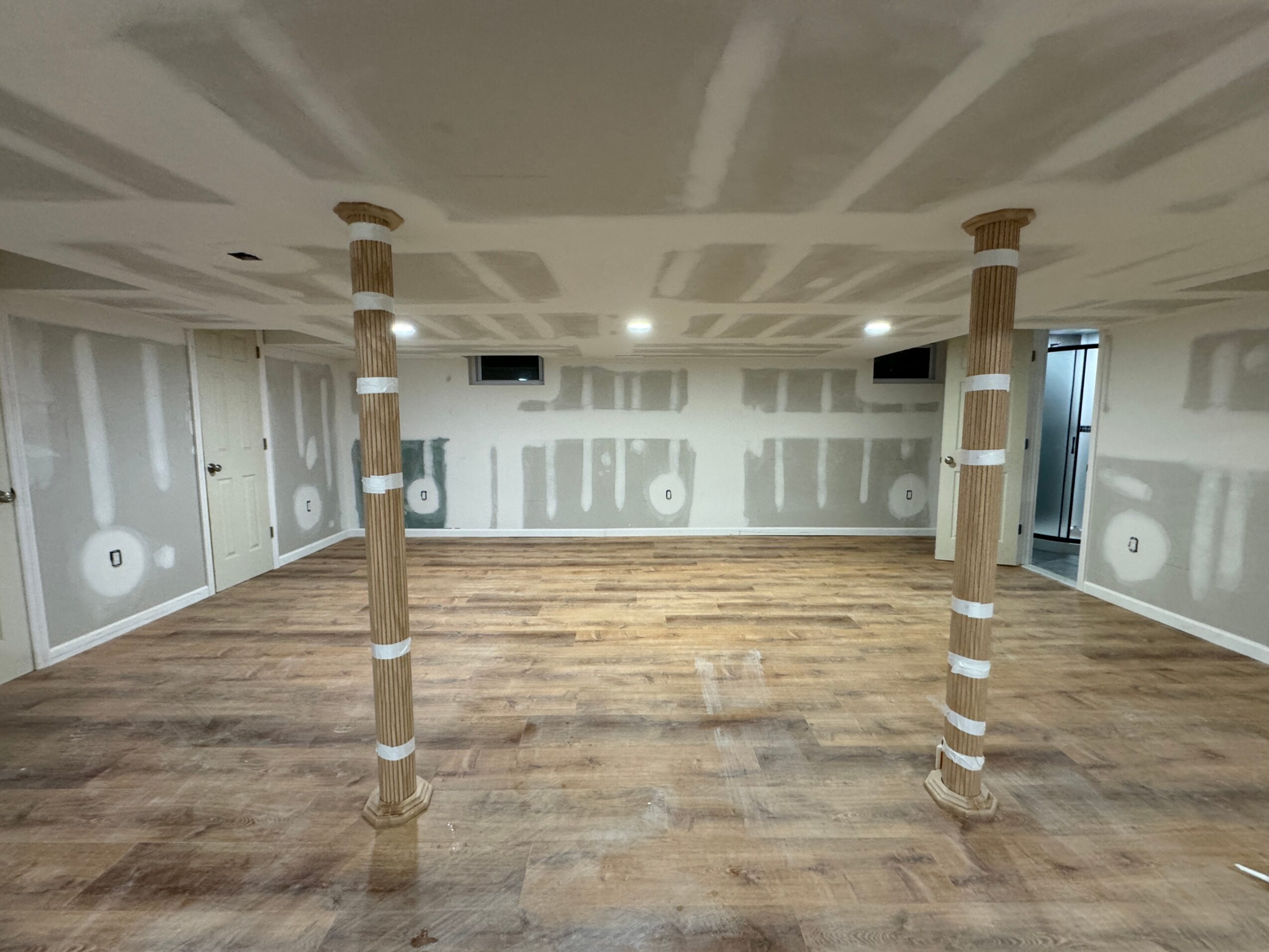 Empty room with unfinished drywall, wooden floor, and two columns in the center, ceiling with visible seams and lighting fixtures.
