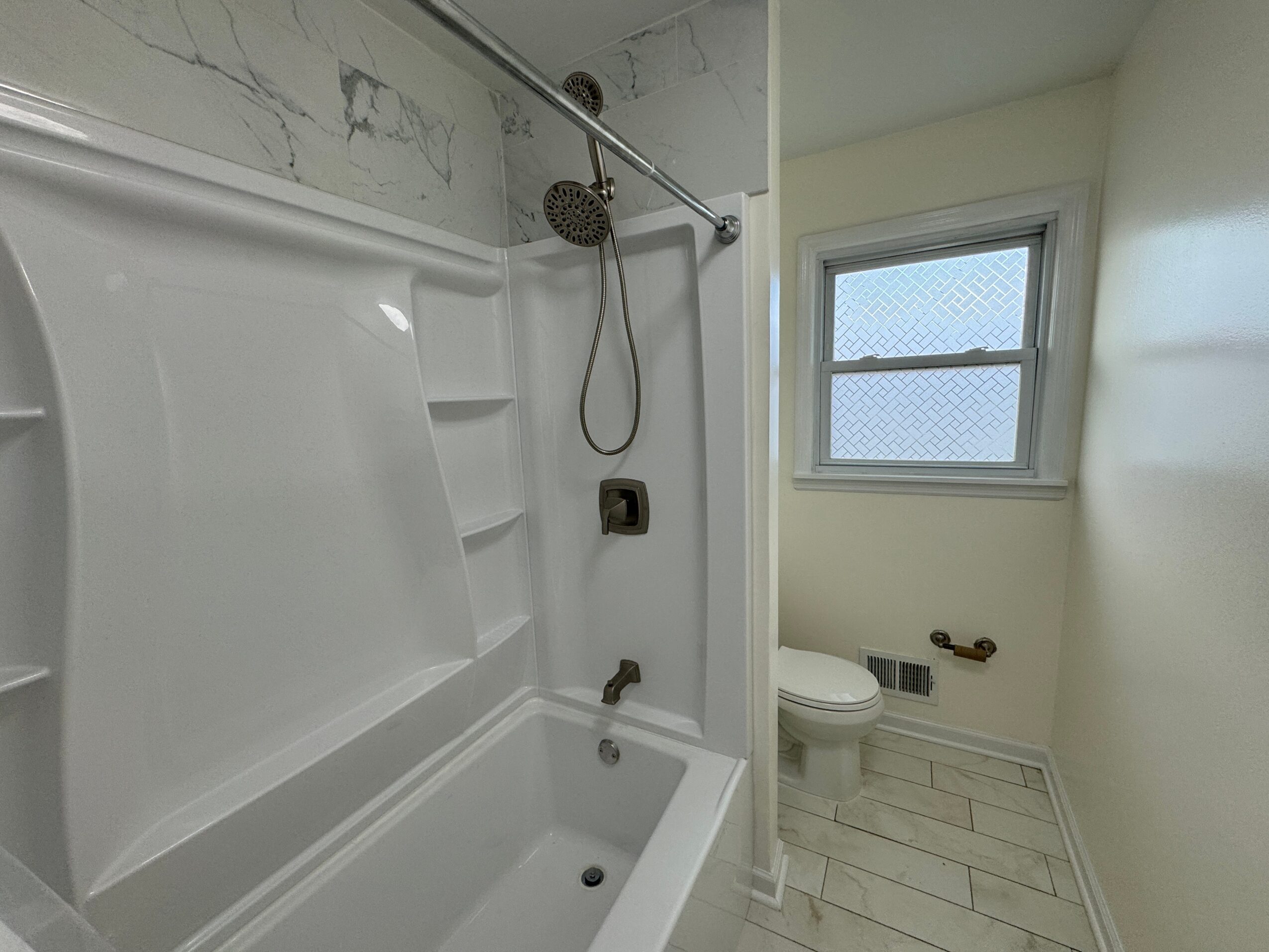 Bathroom with bathtub, showerhead, toilet, and window, with light-colored walls and tiled floor.