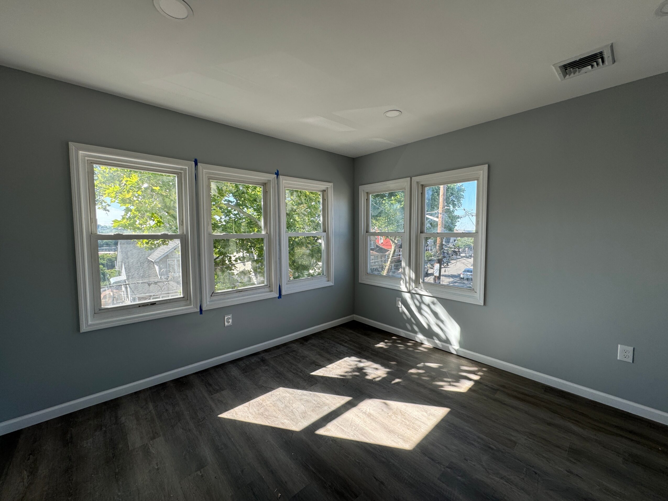 Empty room with light gray walls, four windows, dark wood floor, and sunlight casting shadows inside.