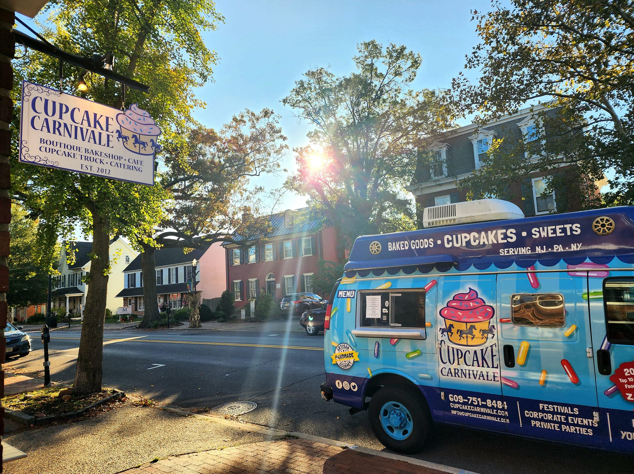 Street scene with colorful cupcake truck, trees, and houses, sunlight shining through trees, and a sign for cupcake carnival.
