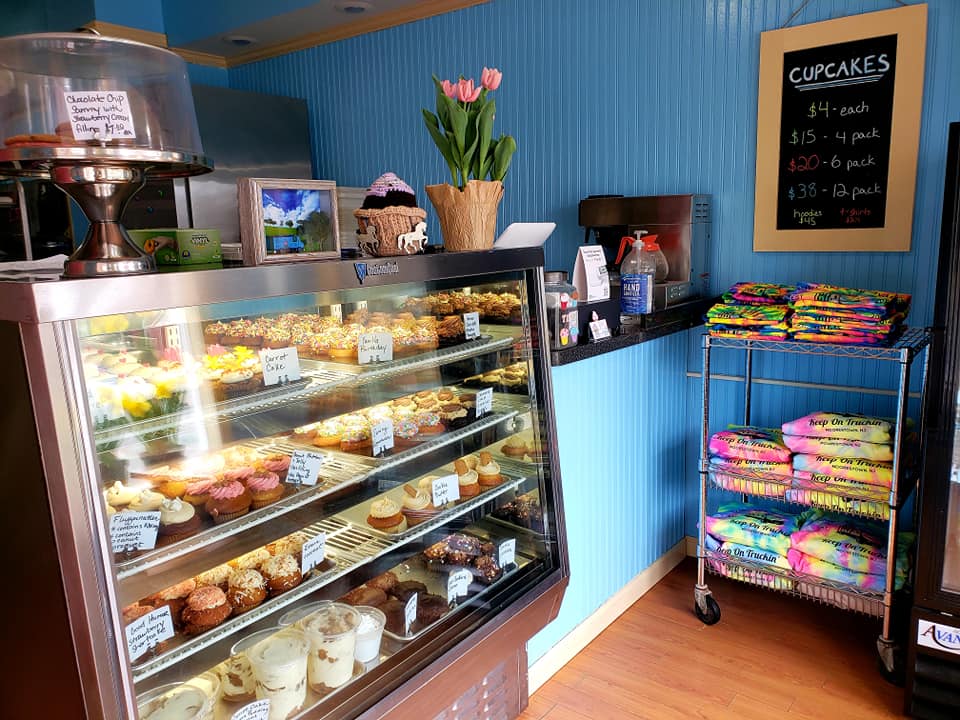 Display case filled with cupcakes, a potted plant with pink flowers, and a chalkboard menu listing cupcake prices.