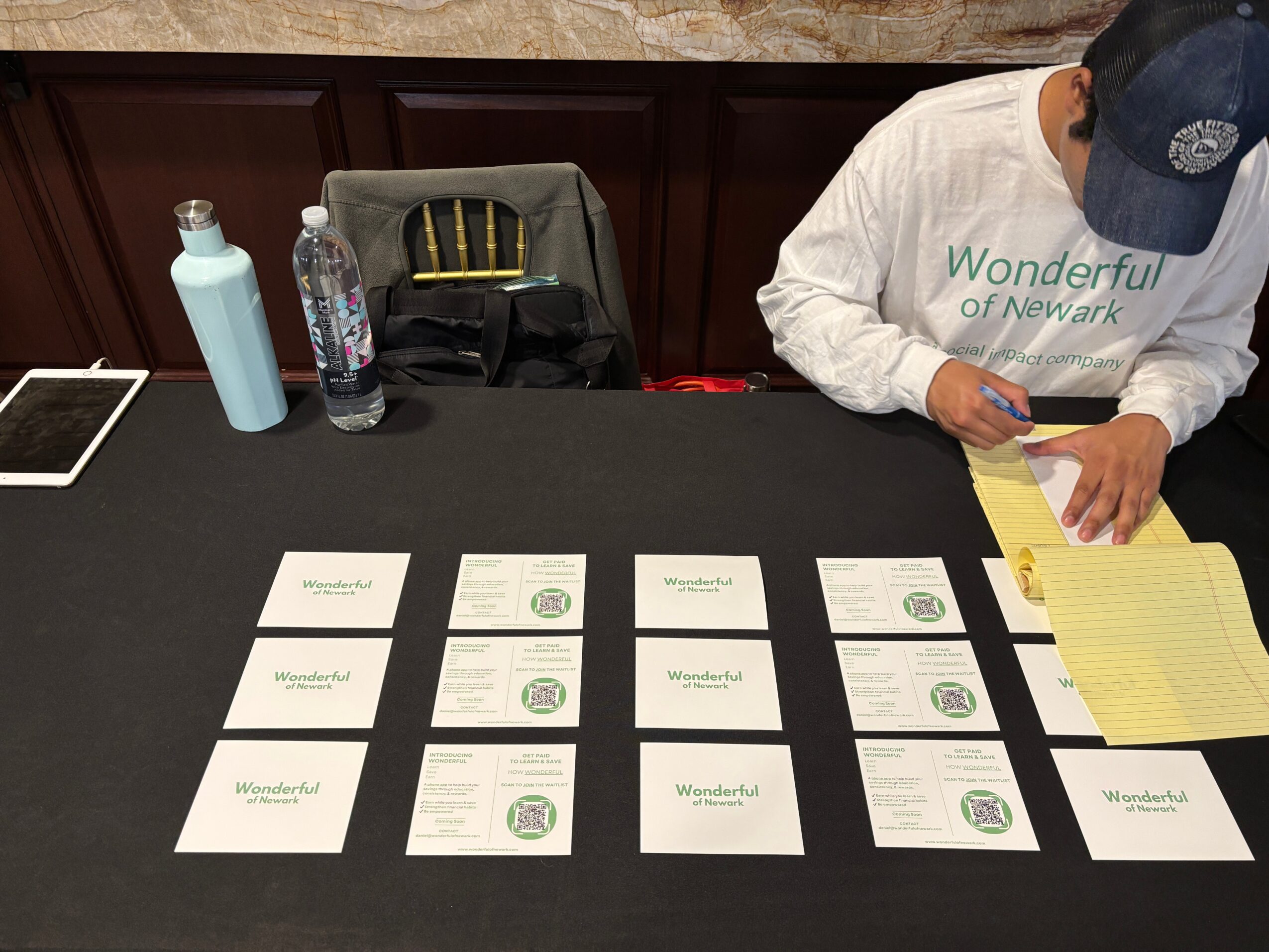 Person writing on yellow notepad at a table with cards, water bottle, and tablet, in a room with dark wood paneling.