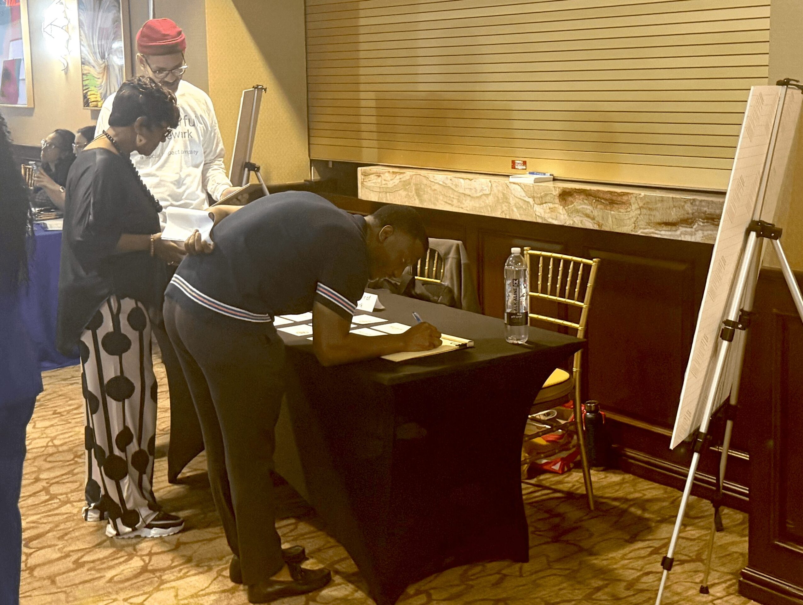Three people gather around a table, one signing a document, in a room with a yellow wall and wooden floor.