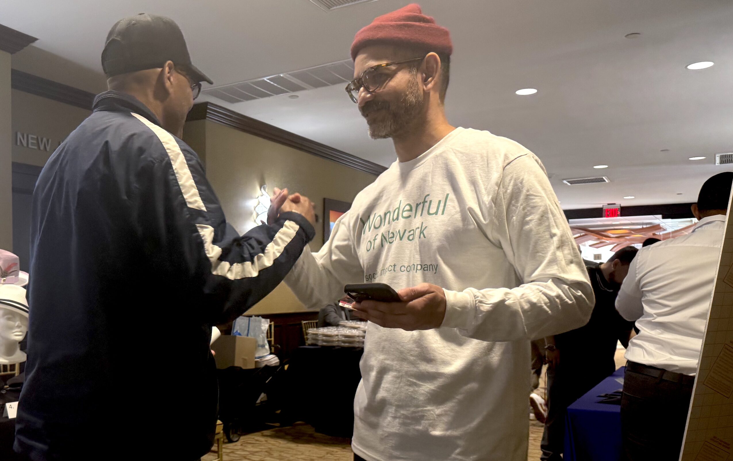 Two men shake hands in a busy indoor setting, one wearing a black jacket and cap, the other in a white sweatshirt and red beanie, holding a phone.