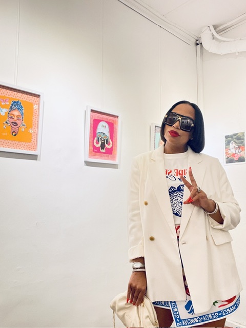 Woman with dark hair, sunglasses, red lipstick, wearing a white coat, standing in art gallery with colorful framed artworks.