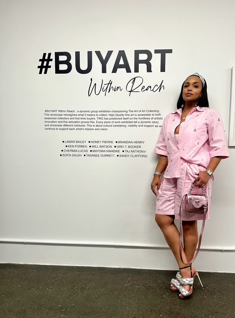 Woman in pink outfit standing against a white wall with text, holding a pink bag, with her right hand on her hip.
