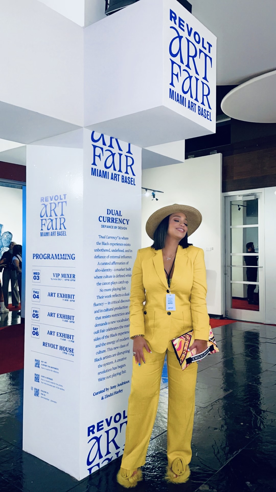 Woman in yellow suit and wide-brimmed hat holding a folder, standing in front of event signage at art fair.