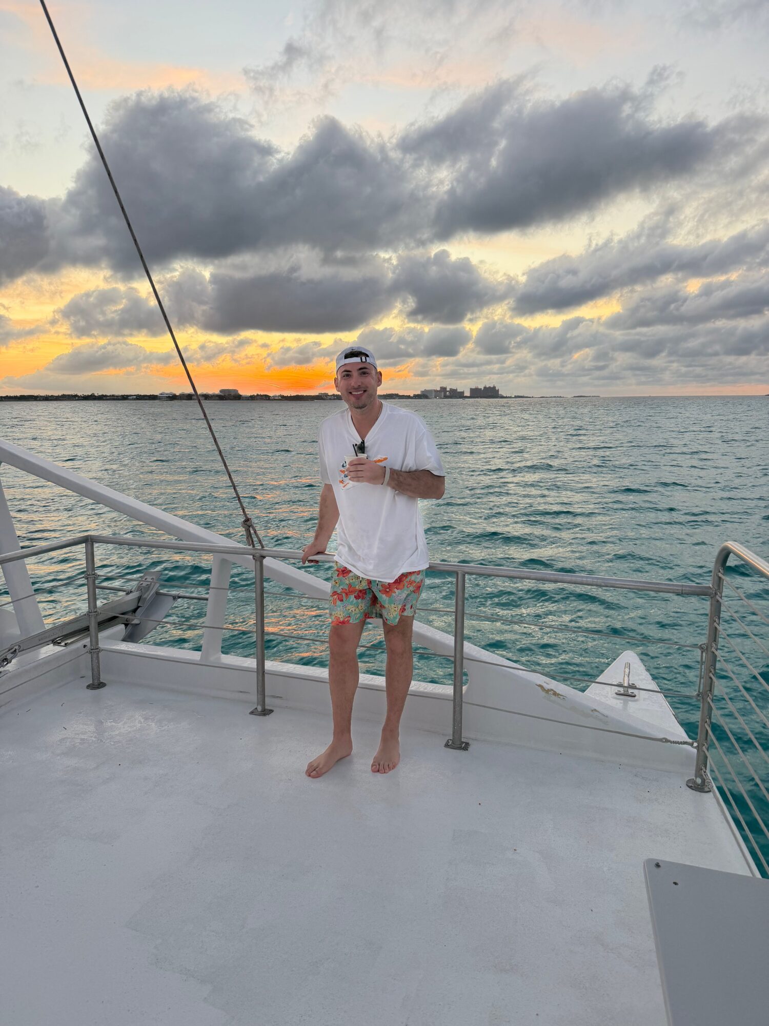 Person standing on boat deck holding a pole, ocean and cloudy sky at sunset in background.