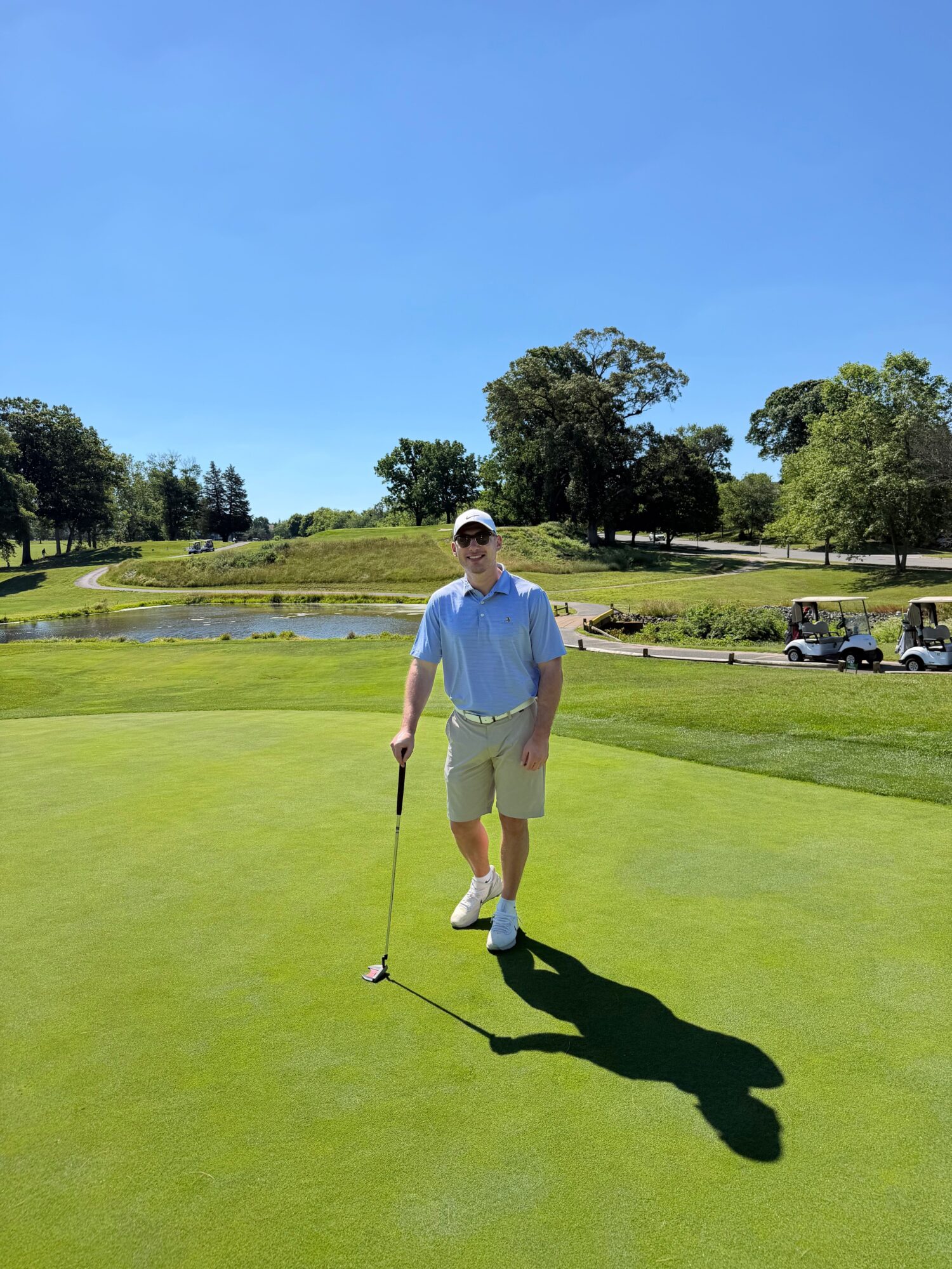 Person standing on a golf course holding a golf club, with a shadow cast on the green grass under a clear blue sky.