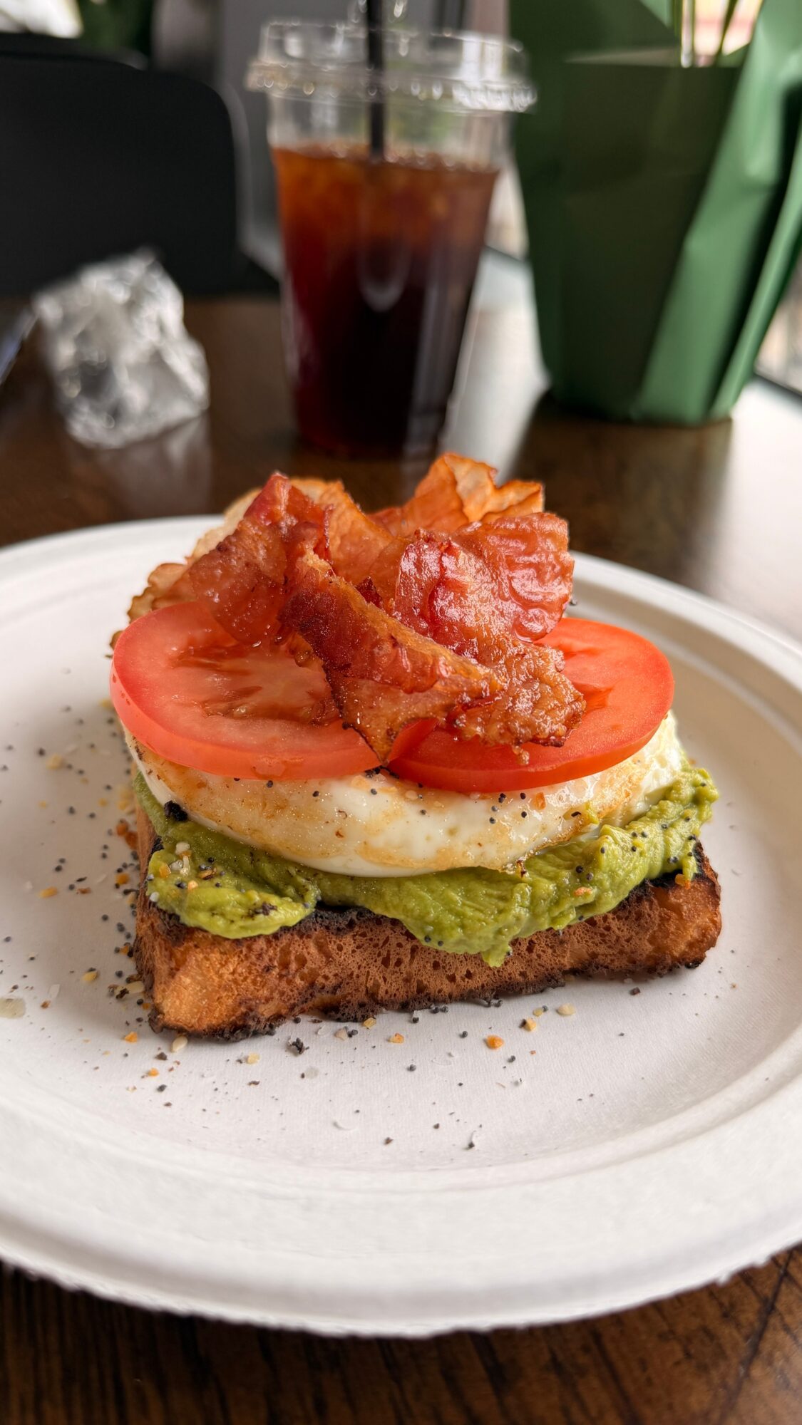 Sandwich with tomato slices, fried bacon, avocado, and fried chicken on a white plate, with a drink and green container in background.