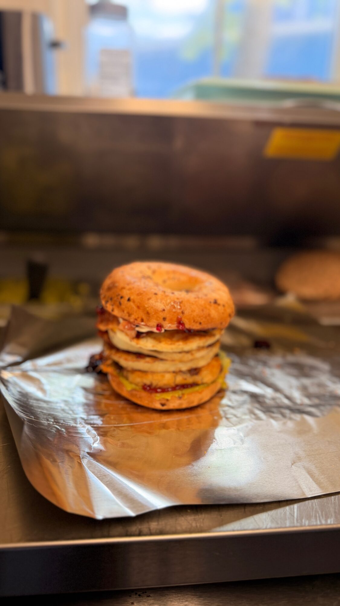 Stacked fried chicken sandwiches with pickles on a foil-lined tray, in a kitchen setting.