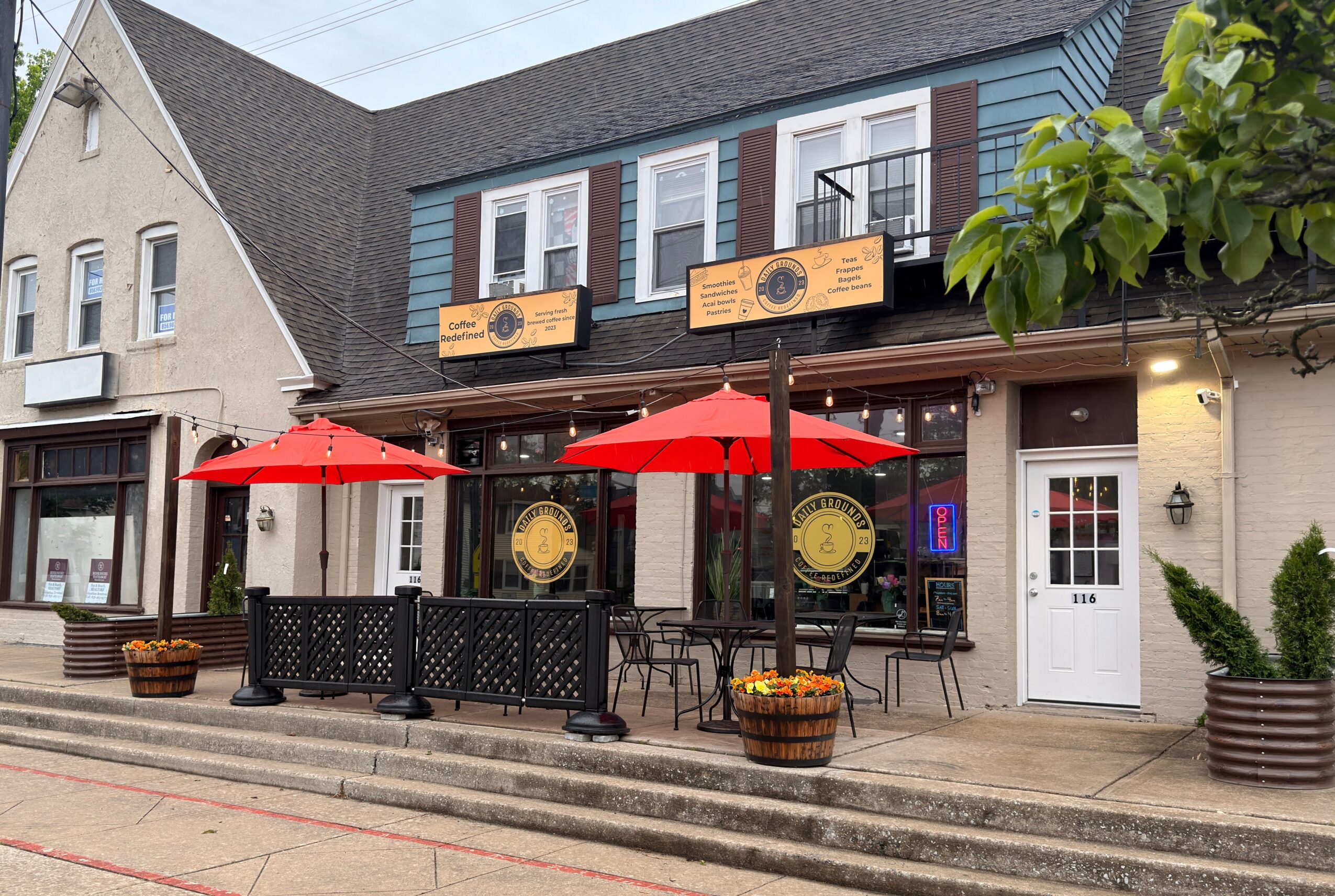 Exterior of a building with outdoor seating, red umbrellas, and potted plants, in a small town setting.